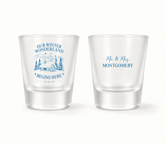 Personalized Our Winter Wonderland Christmas wedding shot glasses featuring snowy mountain cabin artwork, evergreen trees, custom names, and wedding date.