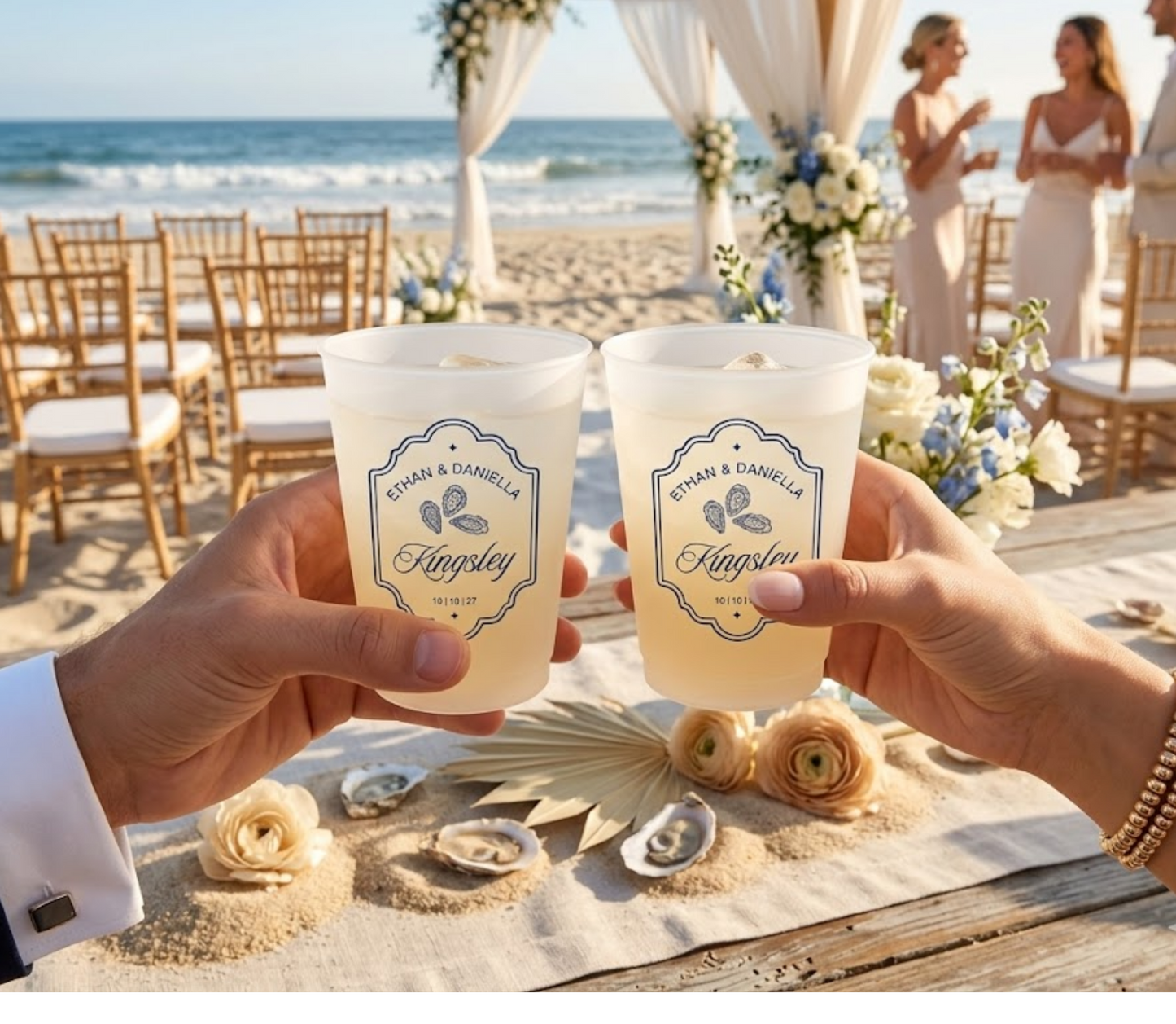 Two hands holding personalized cups at a beach wedding with floral decorations and guests in the background.