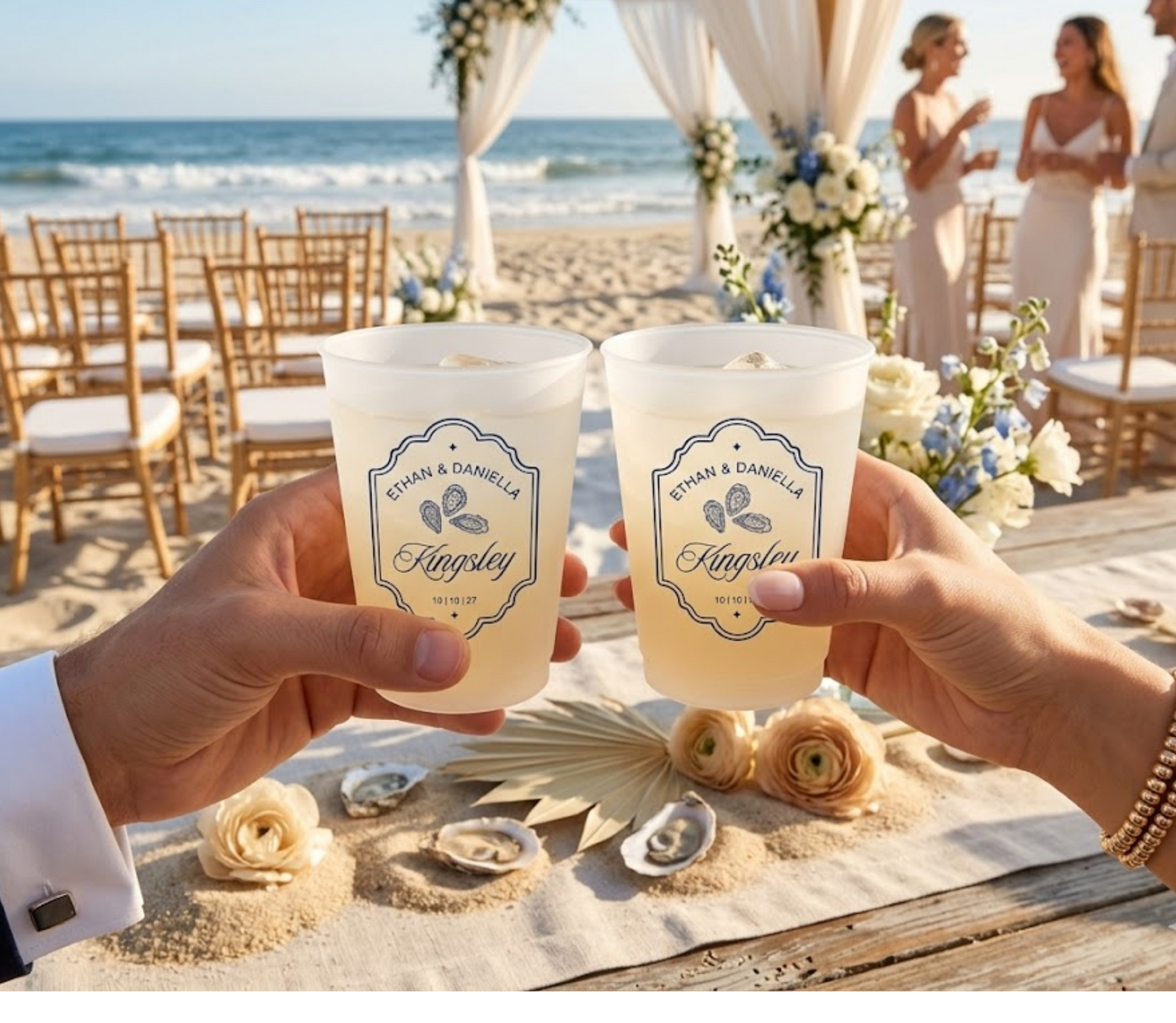 Two hands holding personalized cups at a beach wedding with floral decorations and guests in the background.