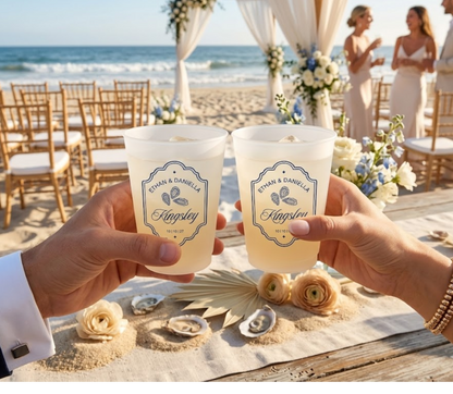 Two hands holding personalized cups at a beach wedding with floral decorations and guests in the background.