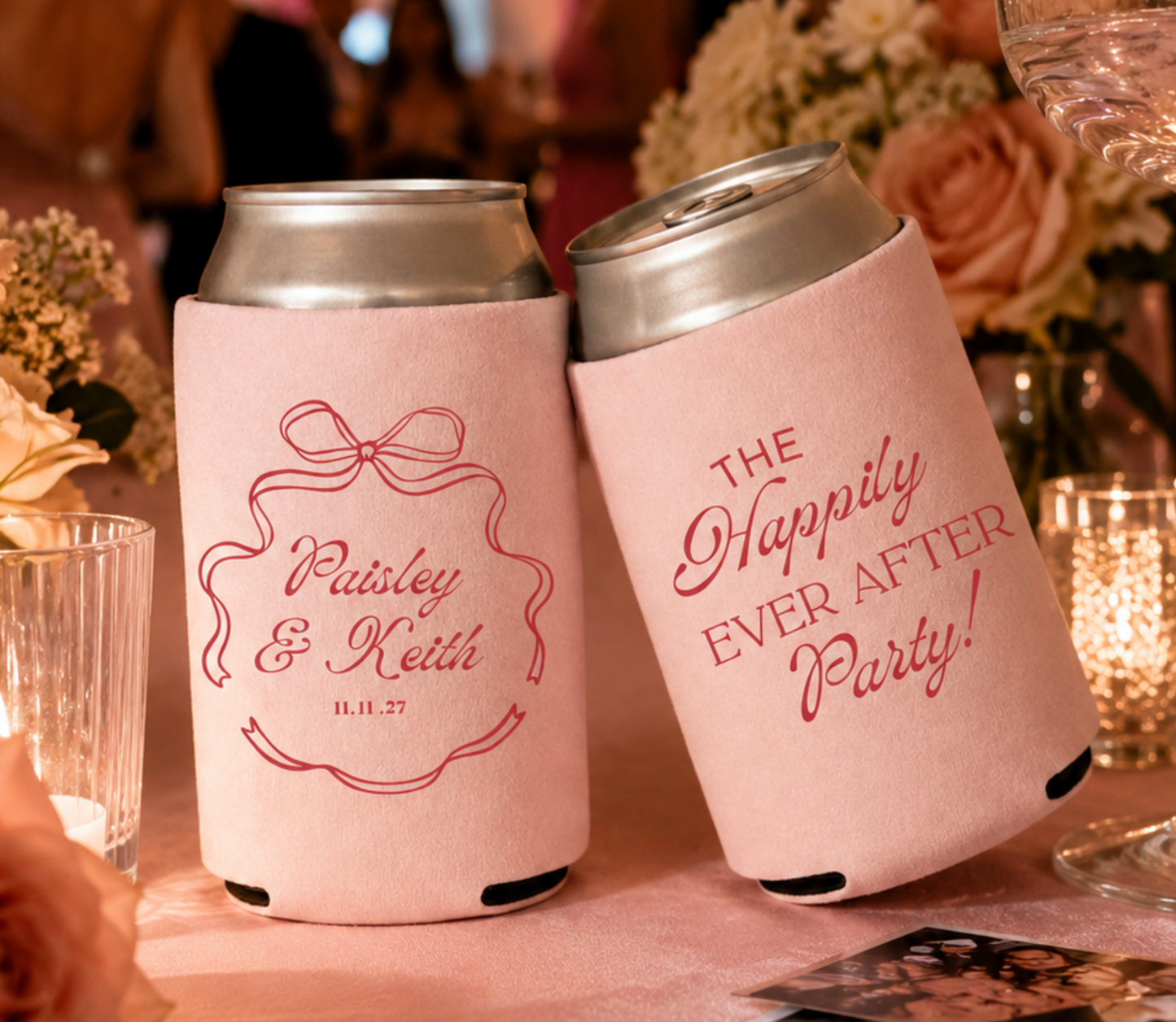 Two pink wedding can coolers with personalized text on a reception table with flowers and glasses.